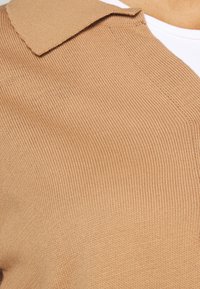 Beige gebreide trui met een V-hals en korte kraag. De textuur is geribbeld met een zachte, lichte stof.