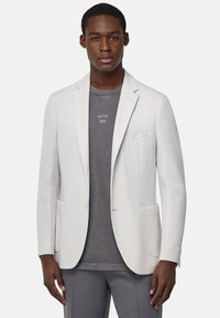 Blazer grigio chiaro in tessuto strutturato con rever a punta, due tasche frontali e chiusura a un bottone, indossato sopra una camicia grigio scuro.