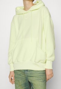 Sudadera de poliéster de color amarillo claro con bolsillo tipo canguro, capucha con cordones y pequeño logo blanco en el pecho, combinada con jeans azul desgastados.