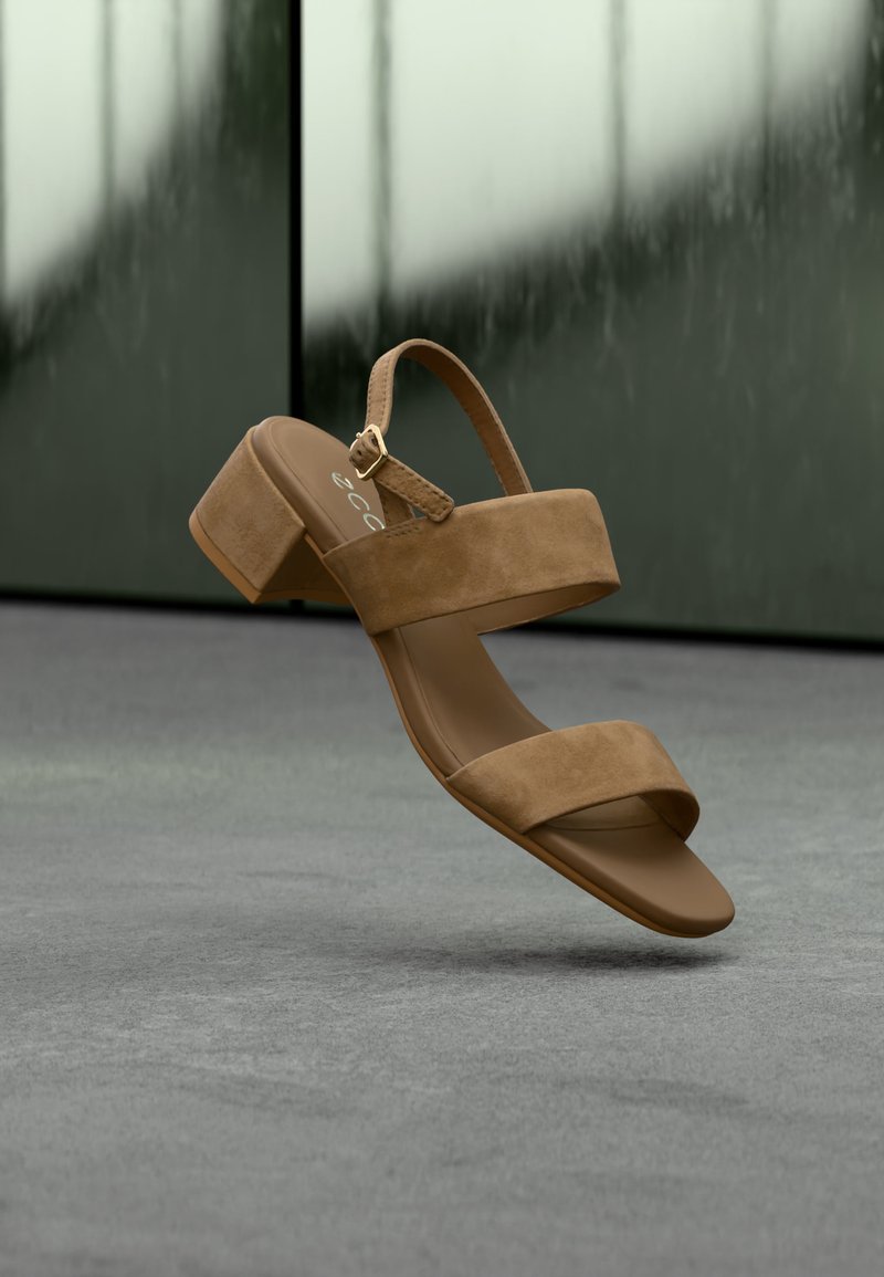 Sandale ouverte en daim marron avec bride à boucle et talon bloc flottant au-dessus d'une surface grise, fond vert foncé flou.