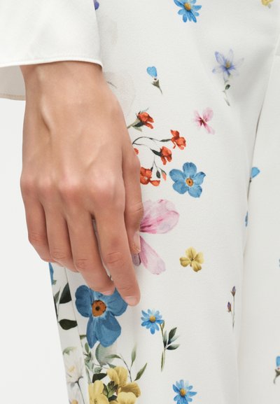 Χέρι που ακουμπά κοντά στο μηρό, φορώντας λευκό ύφασμα με πολύχρωμο ανθισμένο σχέδιο που περιλαμβάνει μπλε, ροζ, κίτρινα και πορτοκαλί λουλούδια.