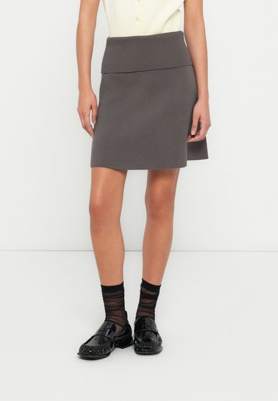 Jupe en maille grise avec une ceinture côtelée, présentant une coupe évasée et une texture douce. Associée à des chaussures noires brillantes et des chaussettes noires à motifs.