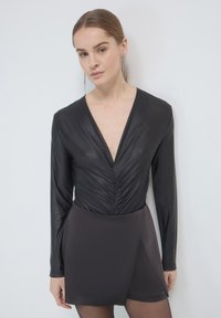 Body nero a maniche lunghe con scollatura profonda a V e dettagli arricciati, abbinato a una gonna pantalone grigia scura con orlo asimmetrico.