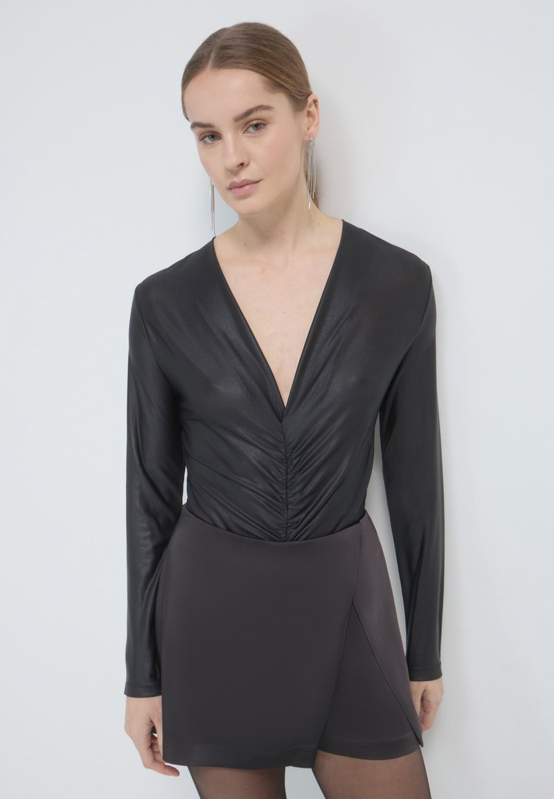 Body nero a maniche lunghe con scollatura profonda a V e dettagli arricciati, abbinato a una gonna pantalone grigia scura con orlo asimmetrico.