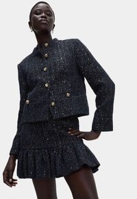 Tkaninowa marynarka w granatowym tweedzie z złotymi guzikami, zestawiona z dopasowaną spódnicą z falbankami. Posiada strukturalny kształt i błyszczące akcenty.