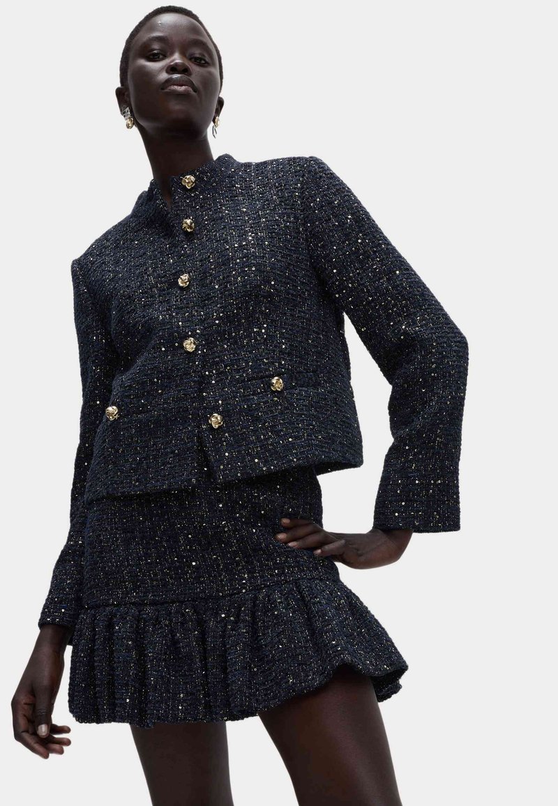 Tkaninowa marynarka w granatowym tweedzie z złotymi guzikami, zestawiona z dopasowaną spódnicą z falbankami. Posiada strukturalny kształt i błyszczące akcenty.