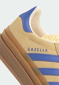 Κοντινό πλάνο μπεζ αθλητικού παπουτσιού Adidas Gazelle με μπλε ρίγες, μπλε ταμπελάκι στη φτέρνα, μπεζ επένδυση και διάφανο λαστιχένιο πέλμα που αποκαλύπτει το λογότυπο της Adidas.