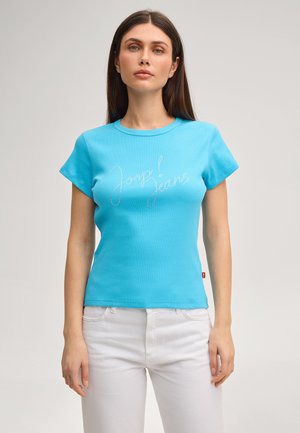 Vrouw draagt een nauwsluitend turquoise T-shirt met de tekst "Joop! Jeans" en witte broek, staand tegen een effen lichte achtergrond.