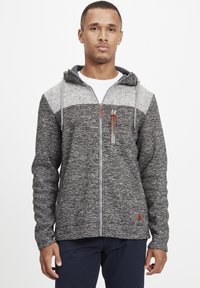 Sudadera con cremallera en gris y negro hecha de un tejido texturizado, que presenta una sección superior de gris más claro, cordones y un pequeño logotipo como detalle.
