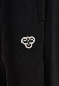 Sort stof med et lille hvidt og sort broderet bi-logo nær en søm, der viser den strikkede tekstur af materialet.