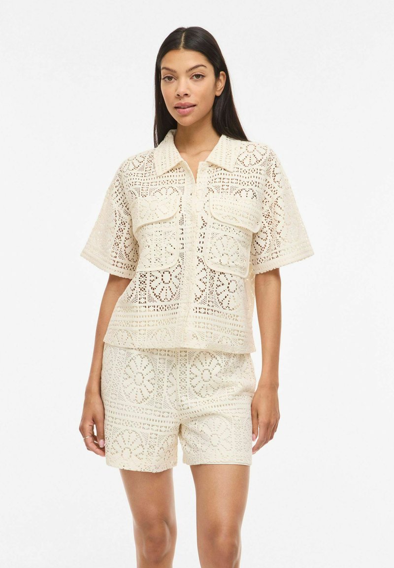 Femme portant une chemise en dentelle crème à manches courtes avec poches avant et un short en dentelle assorti, debout devant un fond blanc.