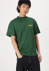 Tricou scurt din bumbac, de culoare verde închis, cu un logo galben pe piept. Guler rotund simplu, croială lejeră și textură netedă.