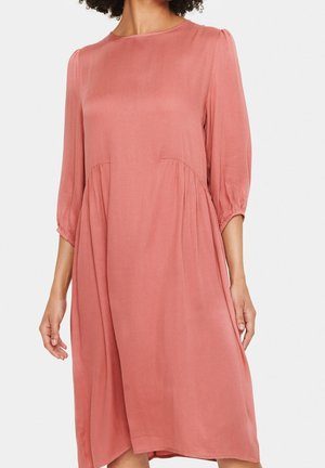 Volně splývavé šaty v délce ke kolenům v barvě prašné růže s kulatým výstřihem a tříčtvrtečními rukávy se shromážděním, které nosí žena s kudrnatými vlasy.