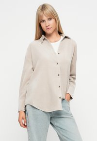 Koszula z długim rękawem w jasnobeżowym materiale, z kołnierzykiem i luźnym krojem. Gładka textura z widocznymi guzikami z przodu.