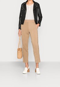 Mujer con una chaqueta de cuero negra, una blusa blanca con un patrón, pantalones beiges, zapatos de tacón bloque blanco y sosteniendo un bolso de paja trenzada.