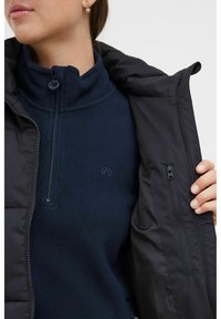 Fleece-pullover i mørkeblå med høy krage og halv glidelås, båret under en svart ytterjakke med glidelomme og en glatt overflate.