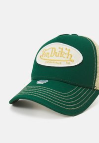 Casquette verte avec un dos en filet beige, ornée d'un patch ovale blanc proéminent avec le logo "Von Dutch Originals" brodé en jaune.