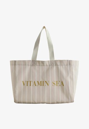 Beige totebag met dunne witte verticale strepen en crèmewitte handvatten, met gouden tekst "VITAMIN SEA" aan de voorkant.
