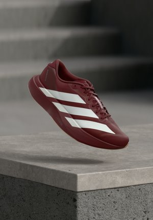 Rood sportschoen met wit ontwerp van drie strepen, lichtgewicht materiaal, gladde textuur en veterluiting, geplaatst bovenop een betonnen oppervlak.