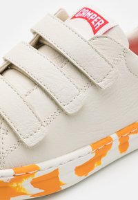 Sneaker bianco in pelle con fasce in velcro, una tomaia testurizzata e una suola dipinta di arancione e bianco. Etichetta del marchio cucita sulla fascia.
