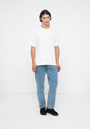 Hvid bomulds t-shirt med korte ærmer, parret med lyseblå denim jeans og sorte sneakers, vist mod en simpel hvid baggrund.