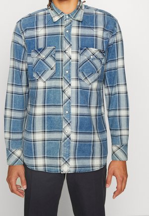 Homme portant une chemise à carreaux bleue et blanche avec deux poches poitrine, associée à un pantalon foncé, debout devant un fond blanc.