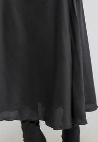 Jupe midi noire, fluide, en tissu léger, avec un ourlet qui descend pour créer une silhouette élégante, associée à des leggings noirs.