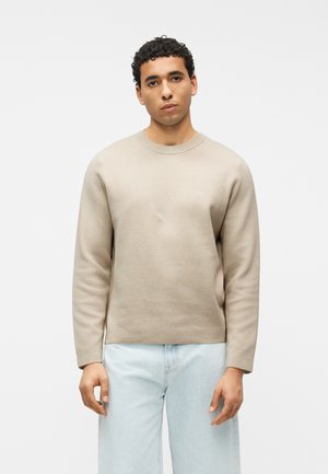 Ung mand med kort krøllet hår iført en beige langærmet sweater og lyseblå jeans, stående mod en ensfarvet hvid baggrund.