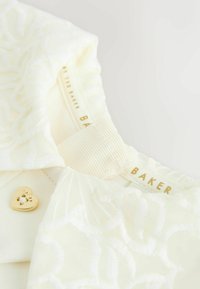 Witte stof met bloemborduursel, gouden hartvormige hardware en een label met de tekst "TED BAKER." Zachte textuur en gedetailleerde stiksels zijn zichtbaar.