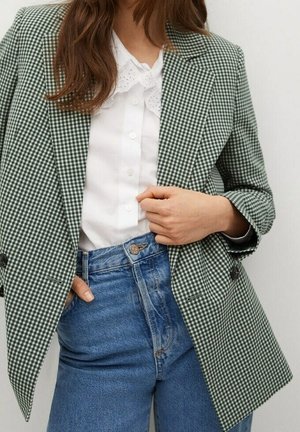 Kvinde iført grøn og hvid ternet blazer, hvid bluse med flæsekant og højtaljede blå jeans med hænderne i lommerne.