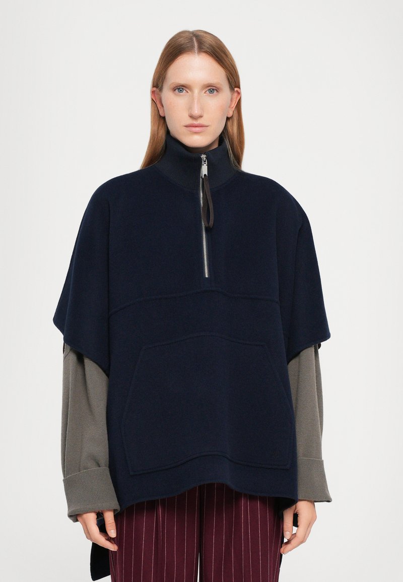 Poncho navy con colletto alto, chiusura a metà zip e tasca frontale. Presenta maniche lunghe grigie a contrasto e polsini a coste.