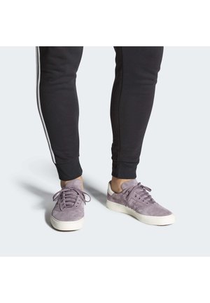 Persona che indossa pantaloni della tuta neri e sneakers in suede viola con suole bianche, in piedi su uno sfondo semplice.