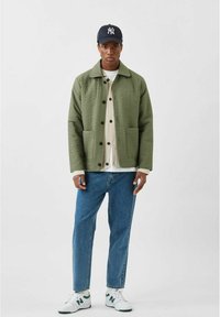 Grøn quiltet jakke med krave, knaplukning og forlommer, lagd over en cremefarvet sweater. Sammen med blå jeans og hvide sneakers.