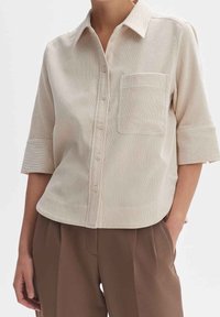 Beige Kurzarmhemd mit Knöpfen und Brusttasche, getragen mit braunen hoch taillierten Faltenhosen, die Hand des Modells in der Tasche.