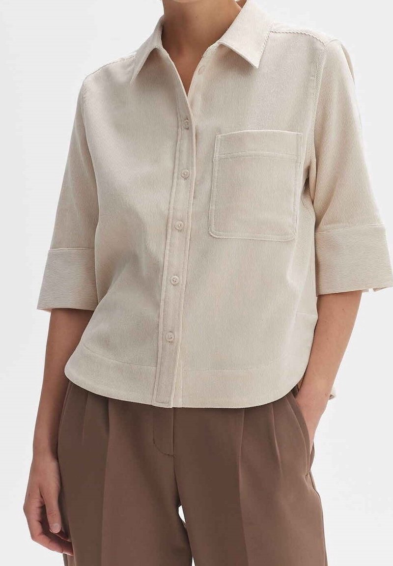 Beige Kurzarmhemd mit Knöpfen und Brusttasche, getragen mit braunen hoch taillierten Faltenhosen, die Hand des Modells in der Tasche.