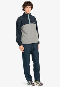 Fleece-pullover i marinblått och grått, med design för halvmåttsdragkedja och grön krage. Kombinerad med marinblå byxor och flerfärgade sneakers.