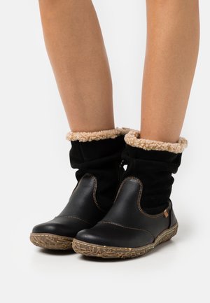 Black leather ankle boots with a fluffy beige collar, textured rubber sole, and decorative stitching along the edges.