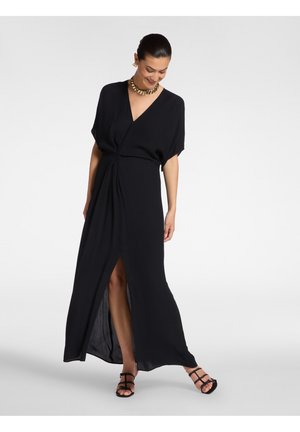 Frau in einem schwarzen Maxikleid im Wickelstil mit kurzen Ärmeln, vorderem Schlitz, goldener Halskette, Ohrringen und schwarzen Riemchensandalen, die lächelnd nach unten schaut.