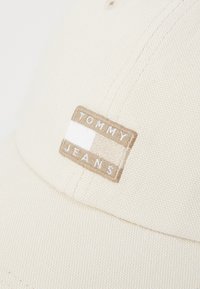 Casquette de baseball beige en tissu texturé, comportant un patch rectangulaire avec "TOMMY" et "JEANS" en lettres blanches.