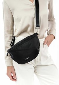 Black nylon fanny pack with a rounded shape, featuring a zip closure and an adjustable strap with a metal clip. Logo patch visible.