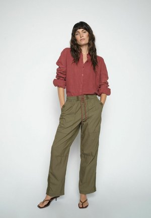 Mujer con cabello oscuro que lleva una camisa roja de manga larga remangada, pantalones verde oliva con cordón y sandalias marrones de tacón, de pie con las manos en los bolsillos.