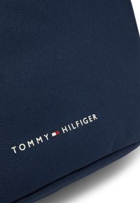 Mörkblått texturerat tyg pryds av "TOMMY HILFIGER"-logotypen i vitt med en liten röd och vit accent. Designen är minimalistisk och modern.