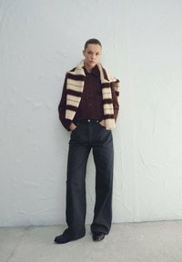 Braune Cordjacke aus Cord mit schwarzen Knöpfen, getragen über einem weiß-braunen gestreiften Schal, kombiniert mit hoch taillierten schwarzen, weit geschnittenen Hosen.