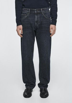 Mann trägt dunkelblaue Jeans mit geradem Bein, schwarze Schuhe und ein dunkles Langarmhemd, steht vor einem einfarbigen hellen Hintergrund.