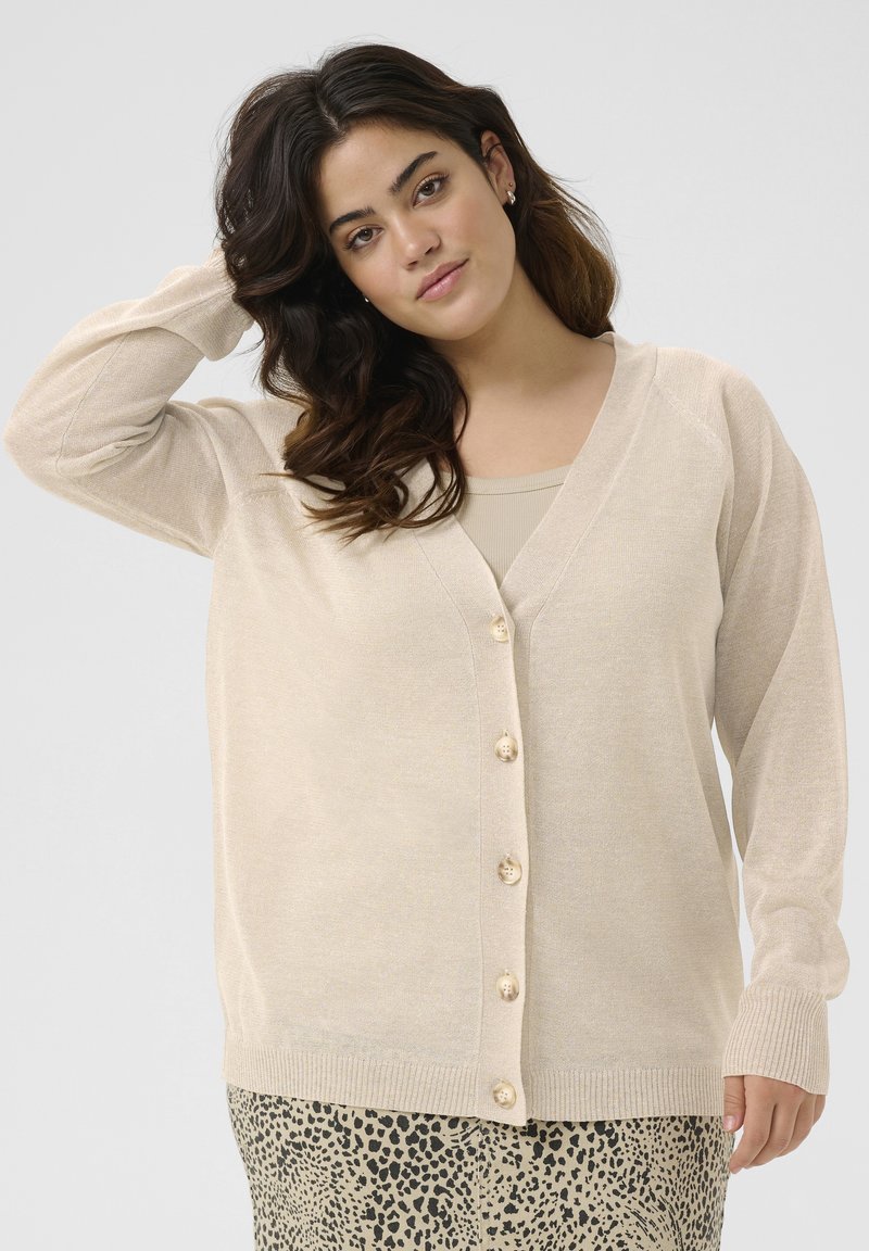 Beige cardigan with a v-neck, long sleeves, and buttons down the front. Soft texture and ribbed hem detail. Worn over a beige top and patterned skirt.