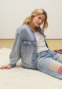 Blonde vrouw met gouden oorringen ligt op een crèmekleurig tapijt, draagt een lichte spijkerjack, een witte blouse en verstelbare jeans.