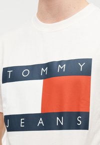 Λευκό βαμβακερό μπλουζάκι με ορθογώνιο σχέδιο λογότυπου σε ναυτικό μπλε, κόκκινο και λευκό χρώμα. Το κείμενο γράφει «TOMMY» και «JEANS» με έντονα γράμματα.