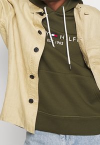 Camisa beige de botones llevada abierta sobre una sudadera verde oliva con cordones blancos y un logo parcial, combinada con unos vaqueros azul claro.