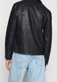 Osoba w czarnej skórzanej kurtce z zamkiem na rękawach i jasnoniebieskich dżinsach, pokazana od tyłu.