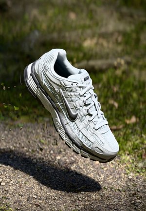 Biały but do biegania Nike w powietrzu nad żwirową ścieżką na tle trawy, pokazujący widok z boku oraz szczegółową podeszwę i sznurówki.